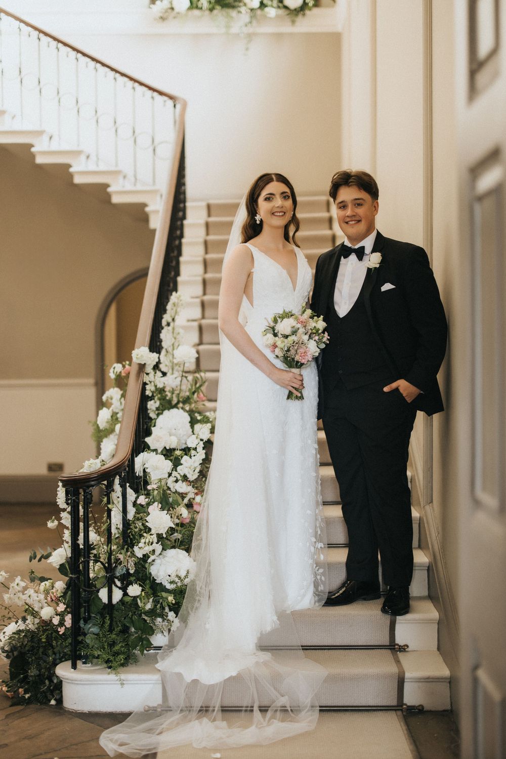 Elegant wedding couple stands on white staircase decorated with white floral arrangements at Saltmarshe Hall - taken by Grimsby photographer