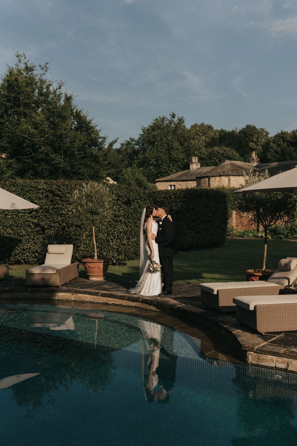 Two figures in elegant attire are reflected in a tranquil blue pool at dusk with outdoor furniture at Saltmarshe Hall.