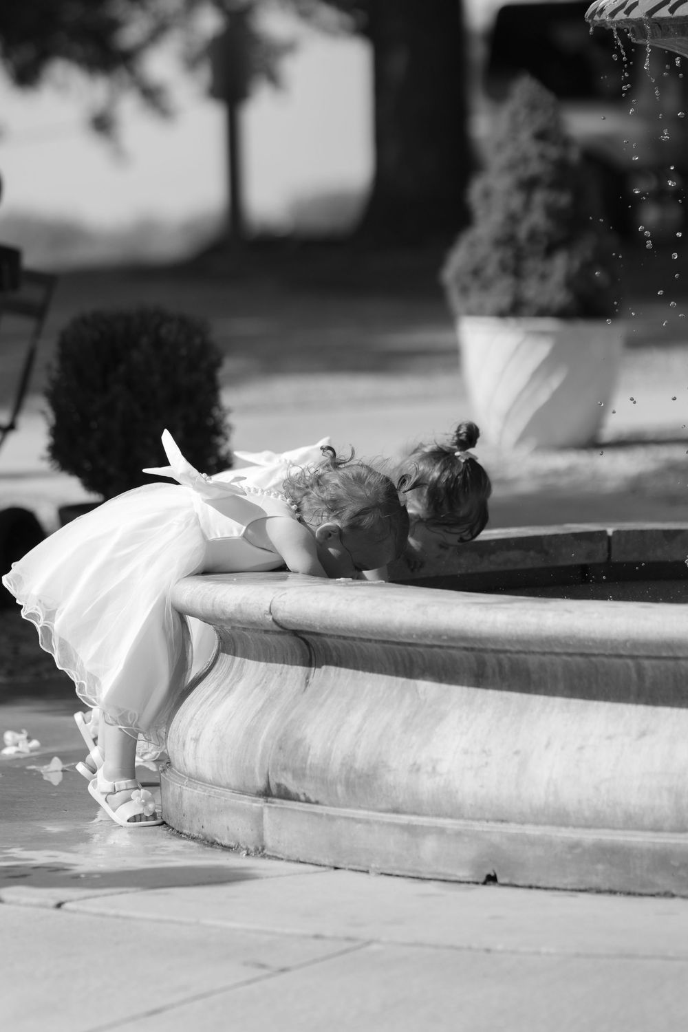 Black and white photo of a young child in white dress looking down into fountain water at the edge.