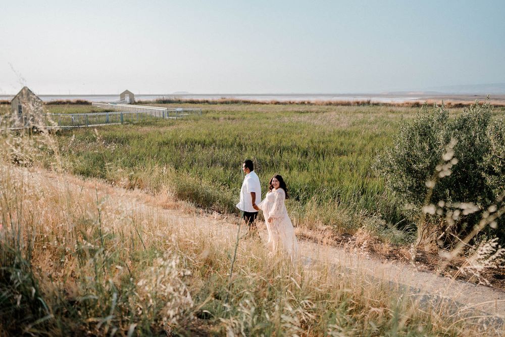 Two figures walk down a winding dirt path through golden grasses and marshland on a sunny day.