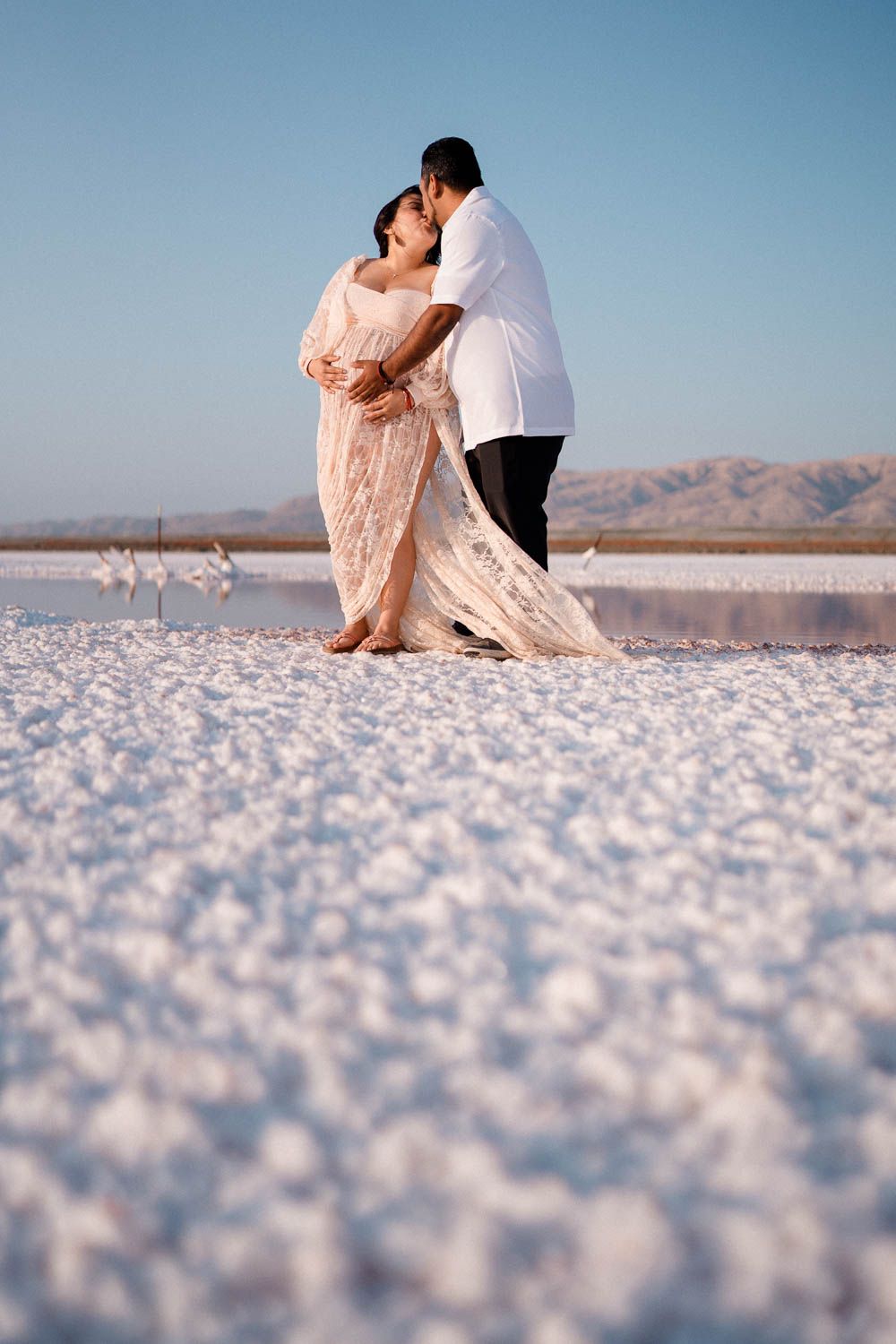A romantic couple embraces on salt flats against distant mountains during a sunset photoshoot.