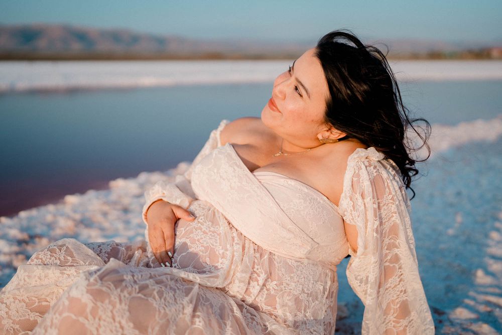 Someone in a flowy lace dress poses gracefully against a tranquil salt lake landscape at sunset.
