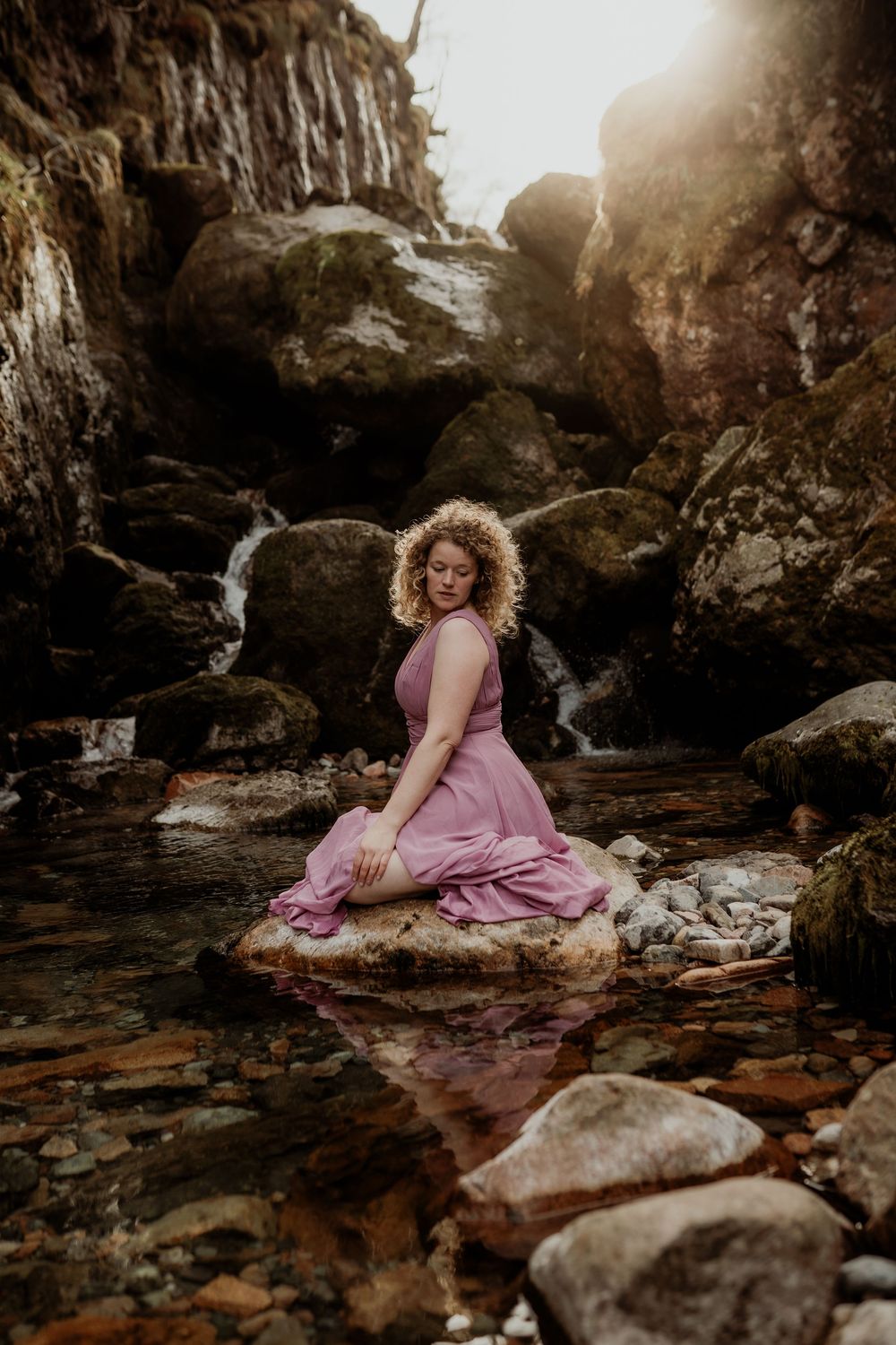 Moody waterfall photoshoot in Glencoe featuring a flowing pink dress reflected in still water beneath rocky outcroppings.
