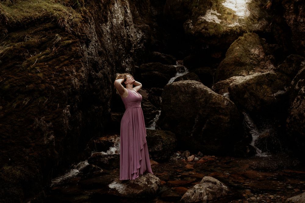 Person in flowing pink dress stands near rocky cliffs and waterfall in moody nature setting.