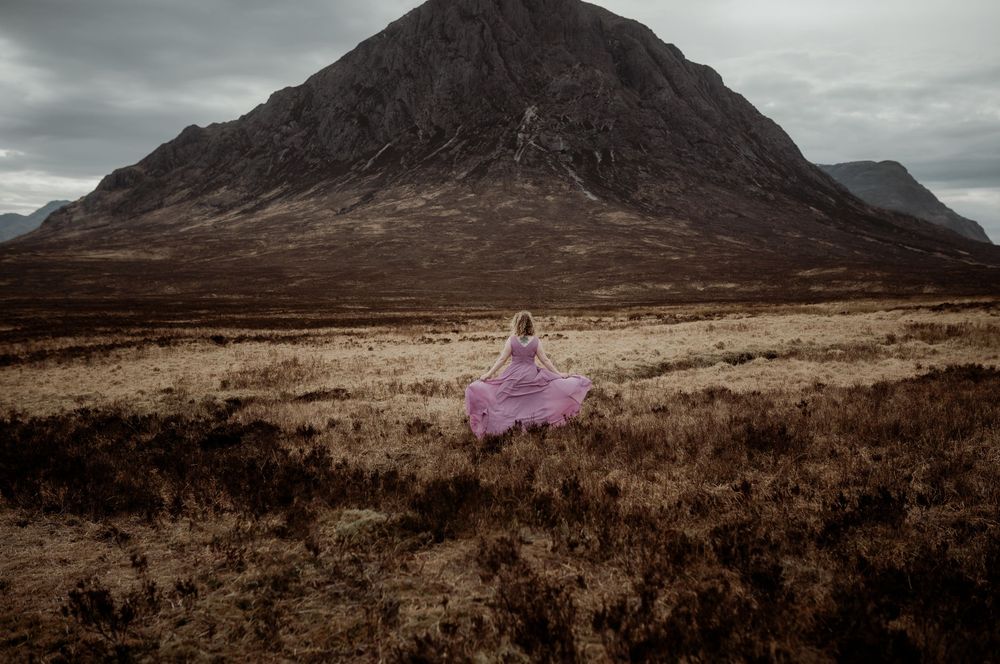 Figure in flowing purple dress stands alone on misty Scottish moorland beneath dramatic mountain peak in moody lighting.