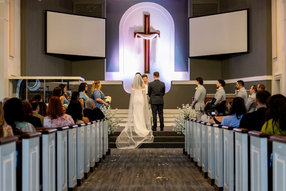Wedding ceremony taking place in modern church sanctuary with illuminated cross altar and white pews filled with guests.