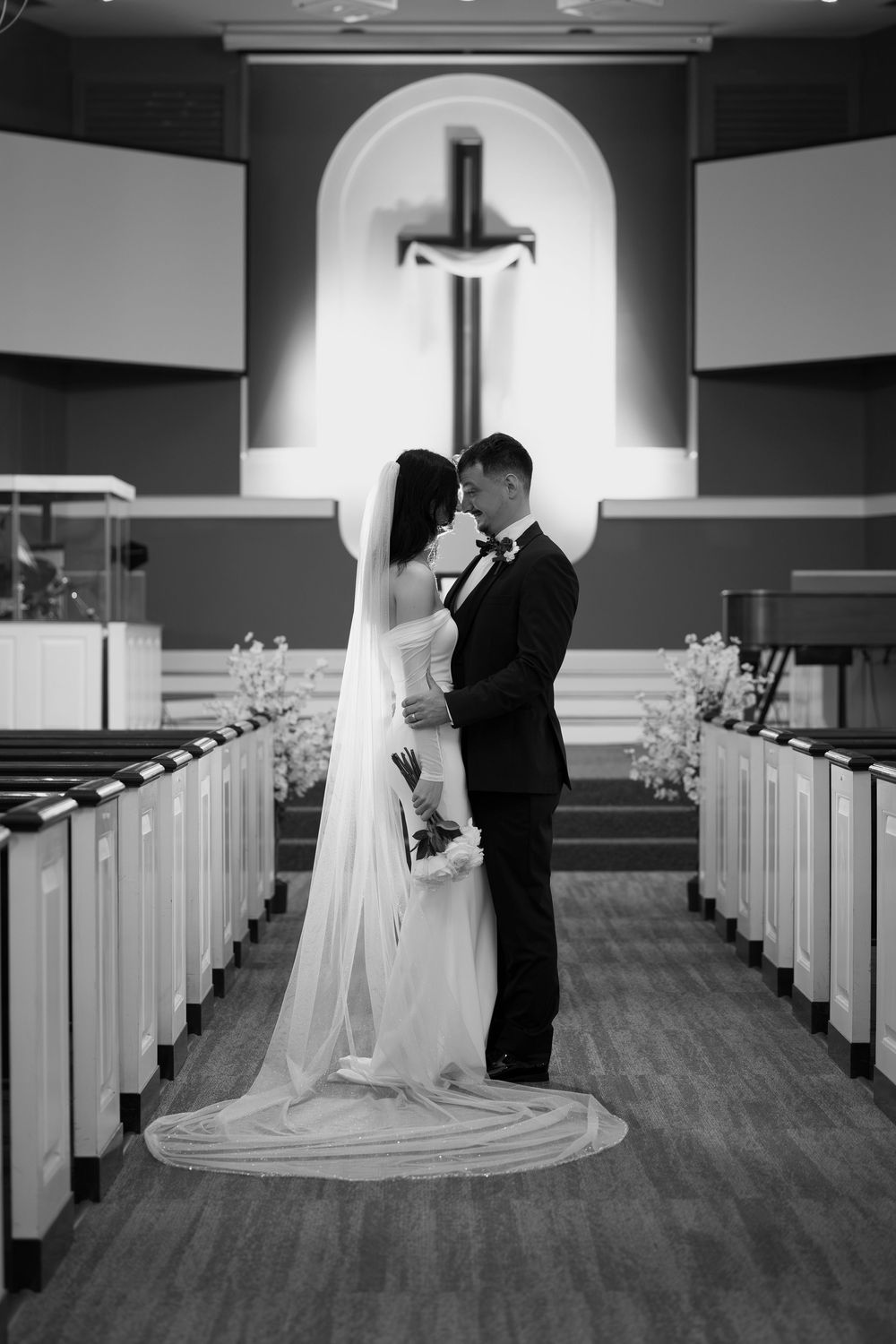 A black and white wedding photo in a church with a cross illuminated in an arched alcove behind the couple.