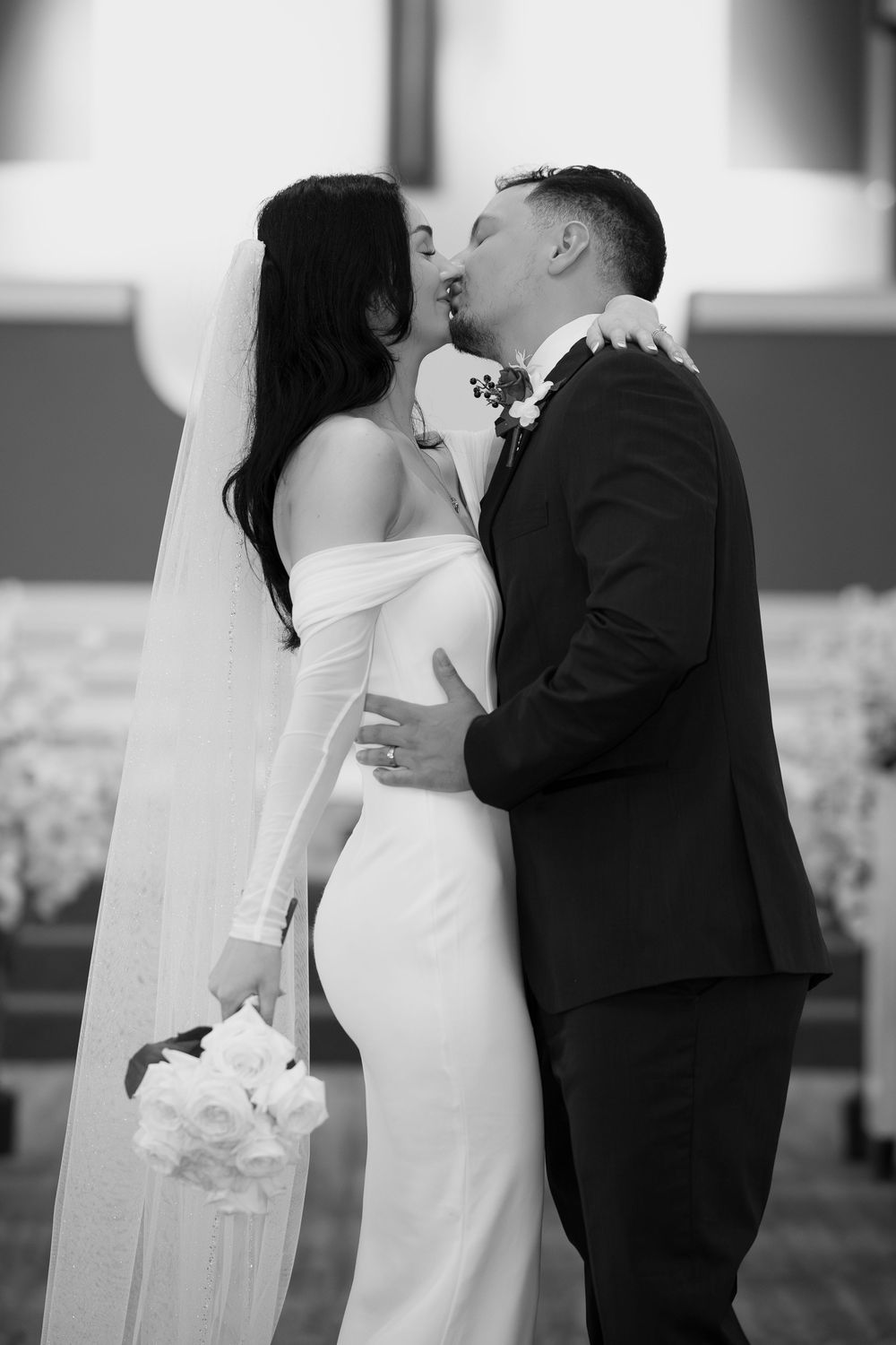 Black and white wedding photograph of newlywed couple kissing while bride holds a white bouquet.