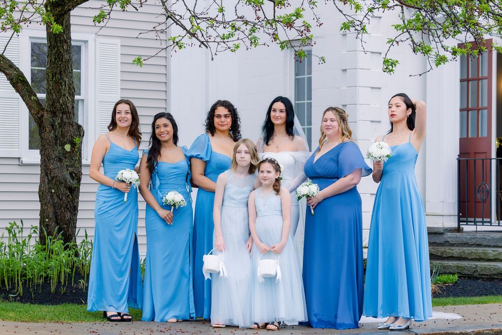 A group of bridesmaids in varying shades of sky blue dresses stand together outside a white church building.