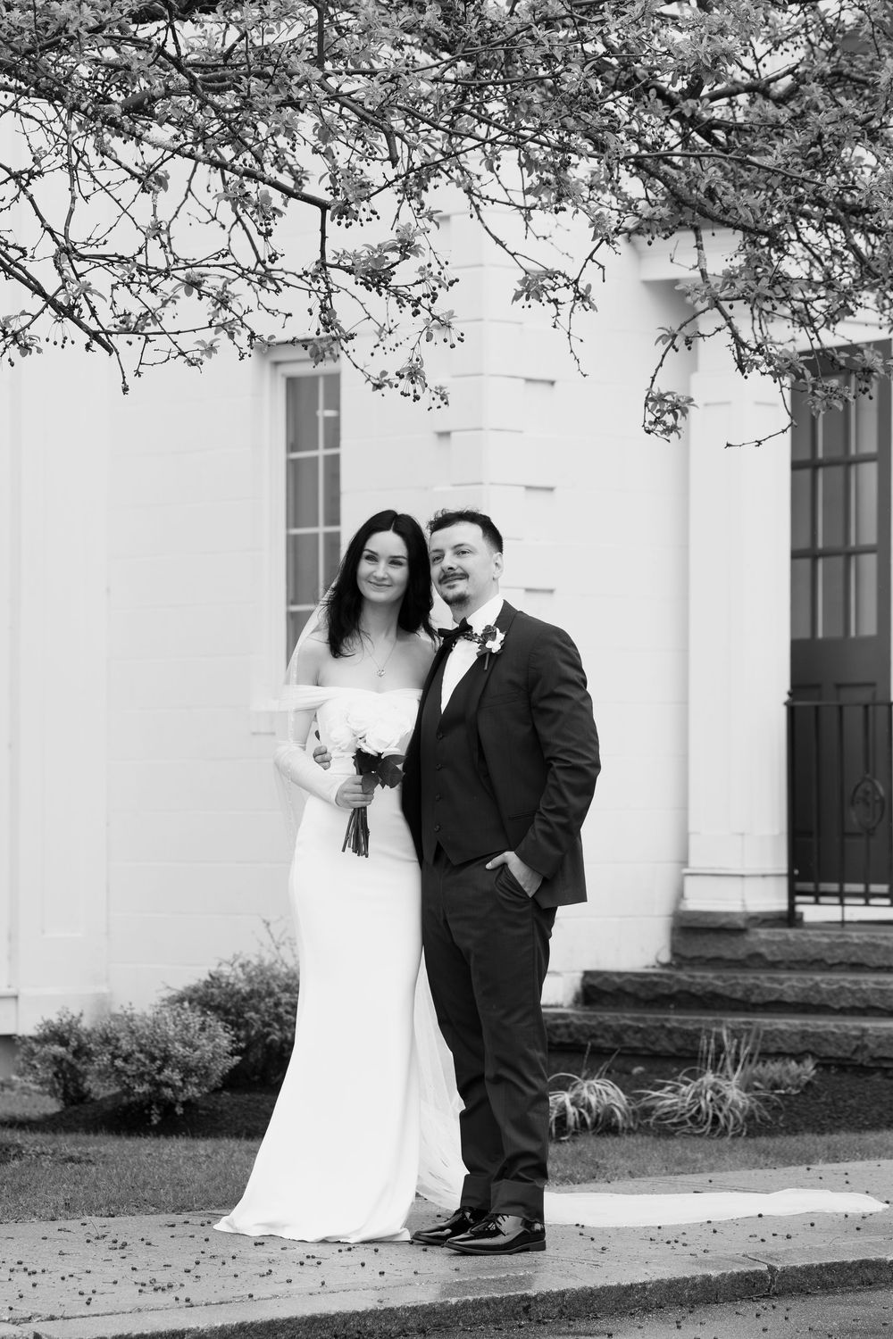 A couple poses together in front of a white colonial-style building with historic architecture in a romantic outdoor setting.