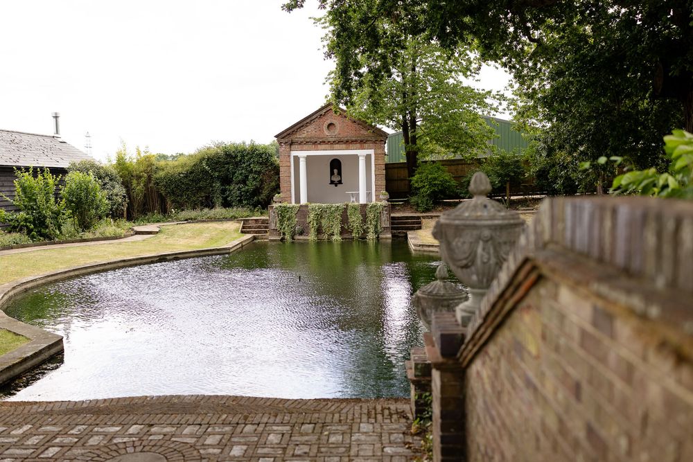 A curved stone pond with a small gazebo structure at one end sits in a tranquil garden setting.