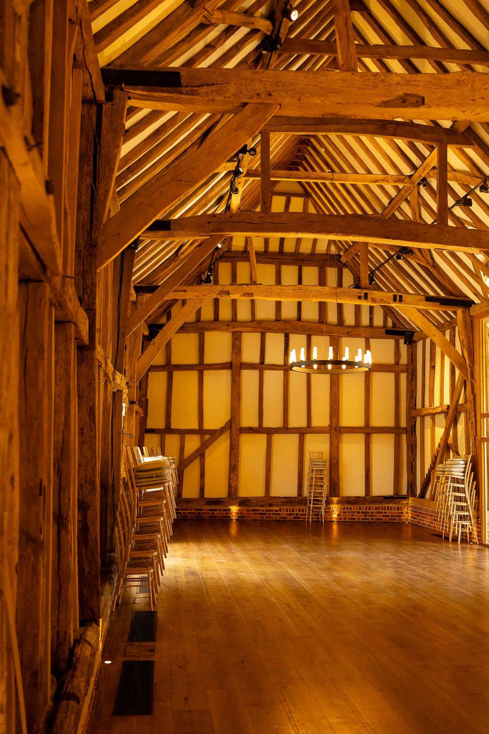 Interior of a rustic barn or hall with exposed wooden beams and hardwood floors illuminated by warm lighting.