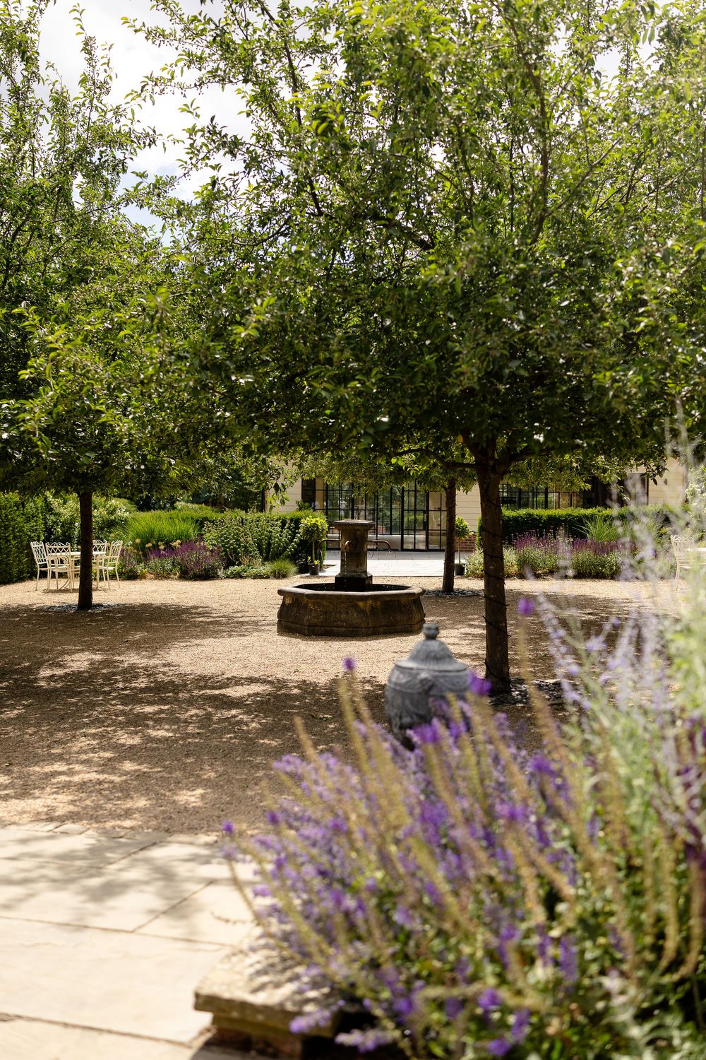 A peaceful garden courtyard with a stone fountain surrounded by tall trees and blooming purple lavender flowers.