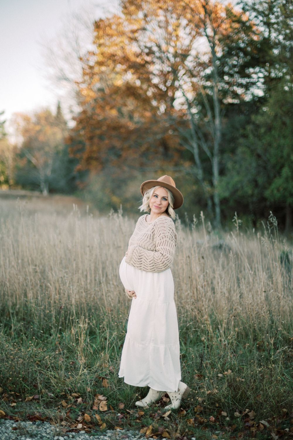 Maternity photo in autumn field showing woman in white dress and beige sweater with brown hat.