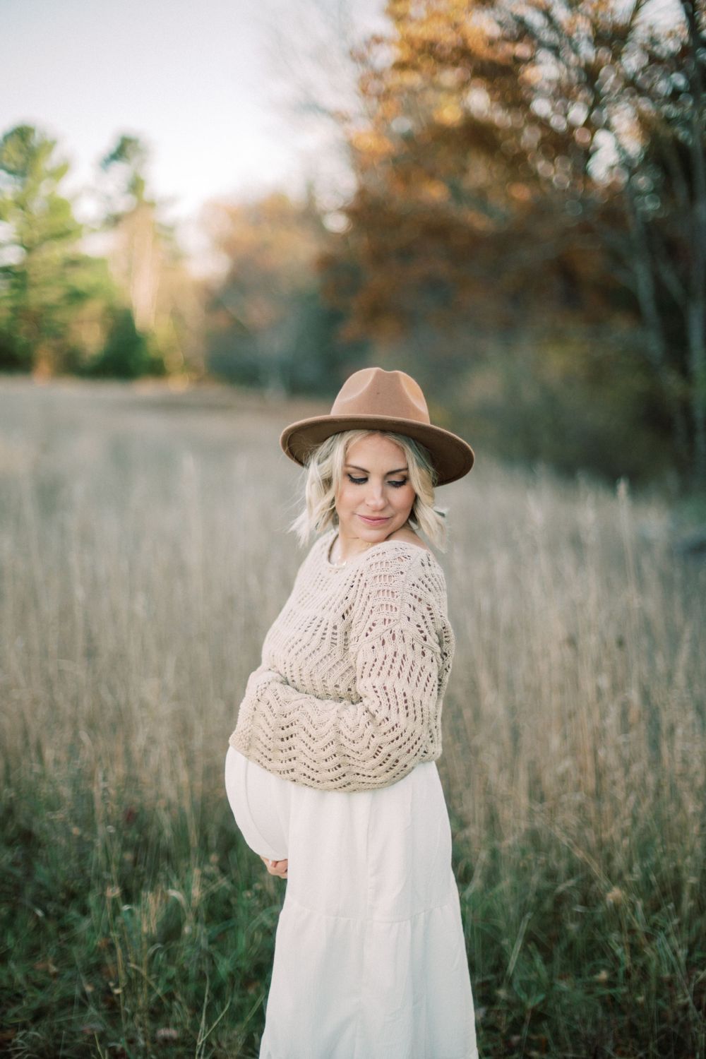 Expectant mother in flowing white dress and knit sweater poses in golden autumn field.