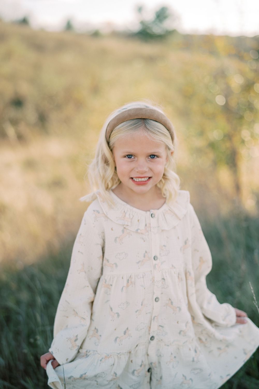 Young girl wearing white dress and headband stands in sunlit meadow.