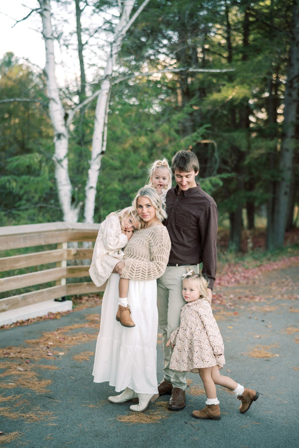 Family poses on wooden footbridge surrounded by autumn trees wearing coordinated neutral outfits.