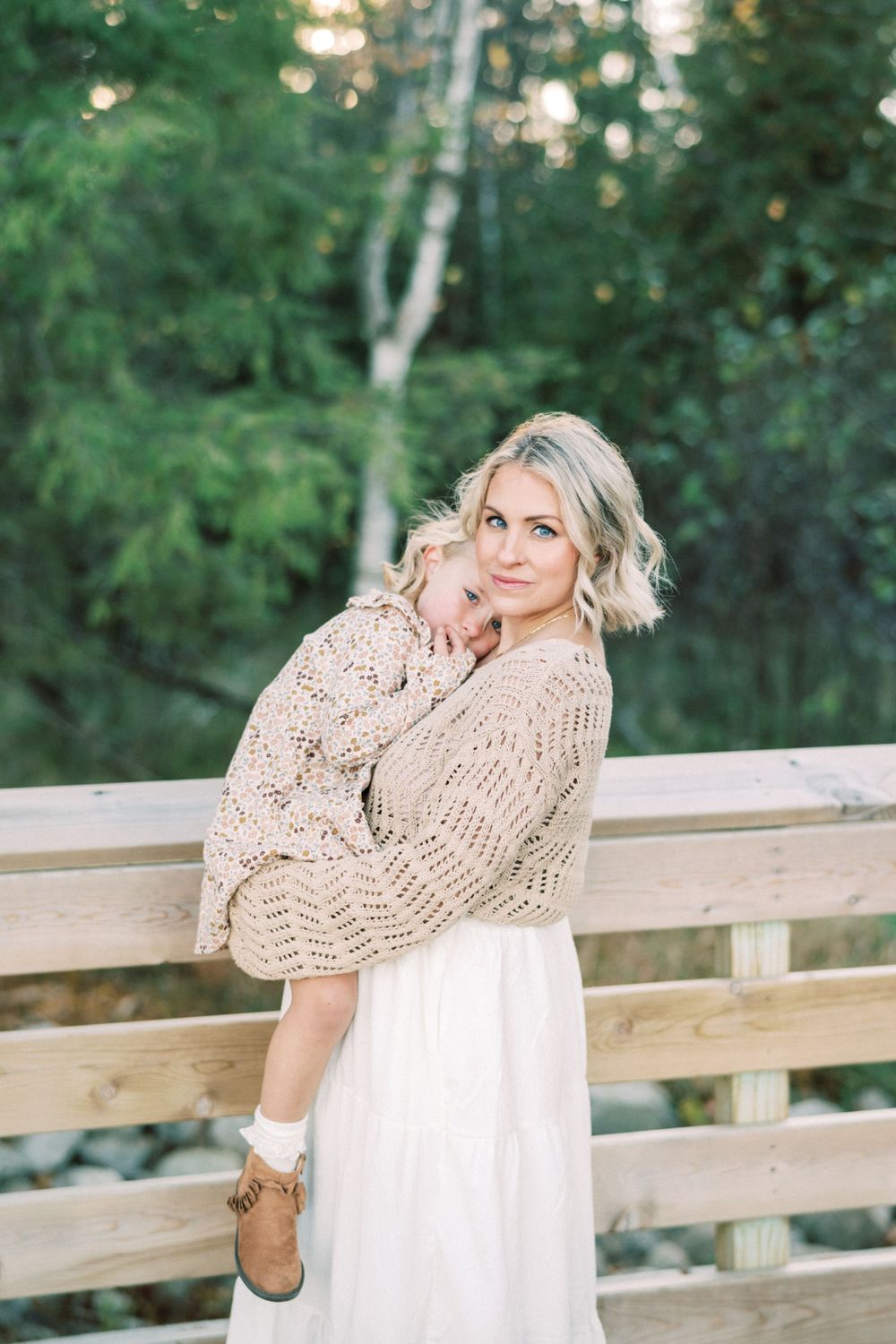 Woman in white dress and beige sweater holds child while posing on wooden bridge deck.
