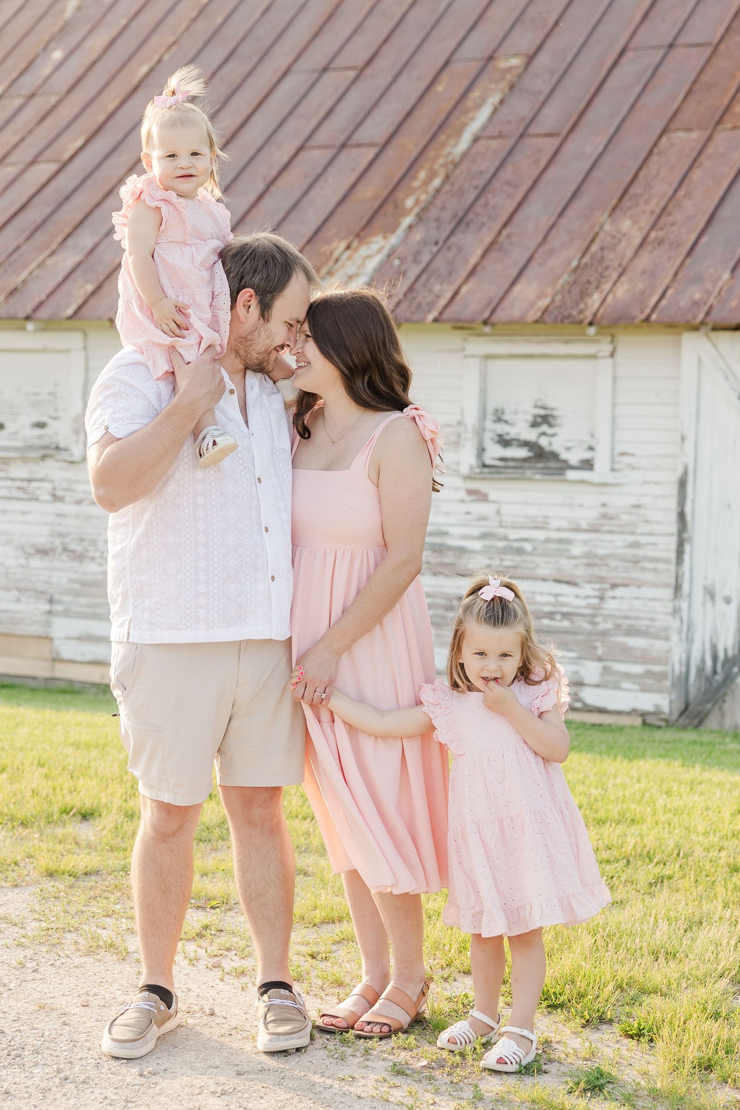 A family wearing coordinated pink outfits poses together outdoors next to a rustic white barn on a sunny day.