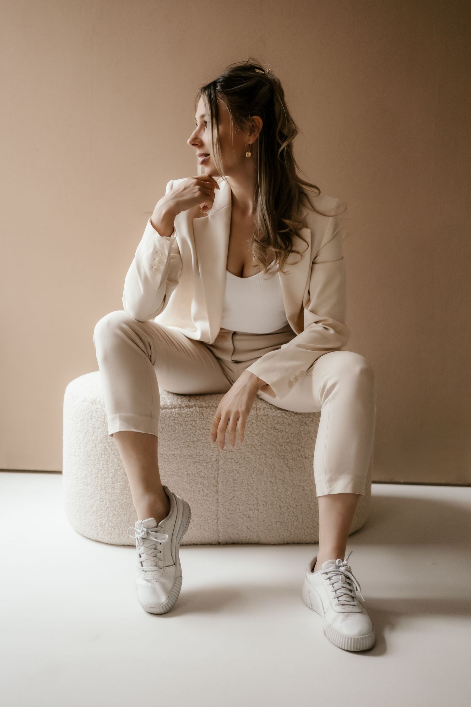 Side profile stylish portrait in monochromatic cream tones showing relaxed pose on textured ottoman wearing white suit and sneakers.