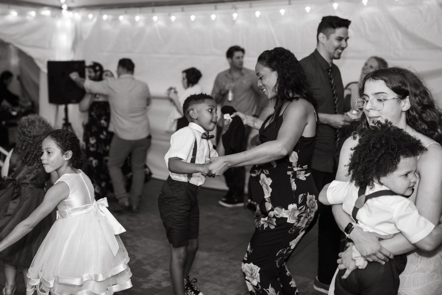 Black and white photo of people dancing and celebrating at a festive event under a tent with string lights.
