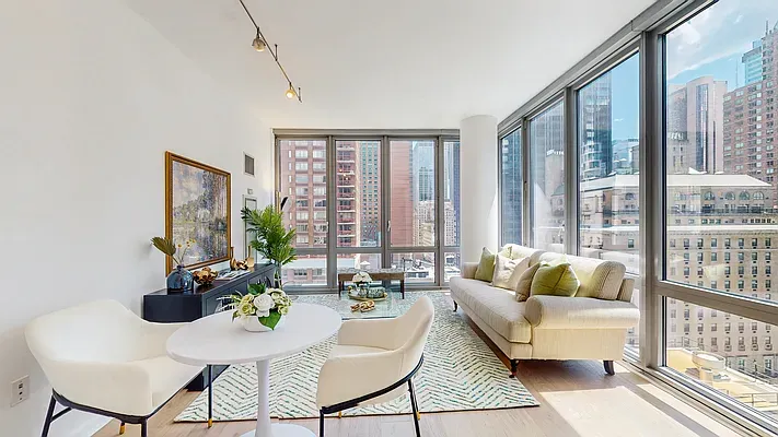 Modern apartment living room with floor-to-ceiling windows, beige sofa, and geometric area rug overlooking city buildings.