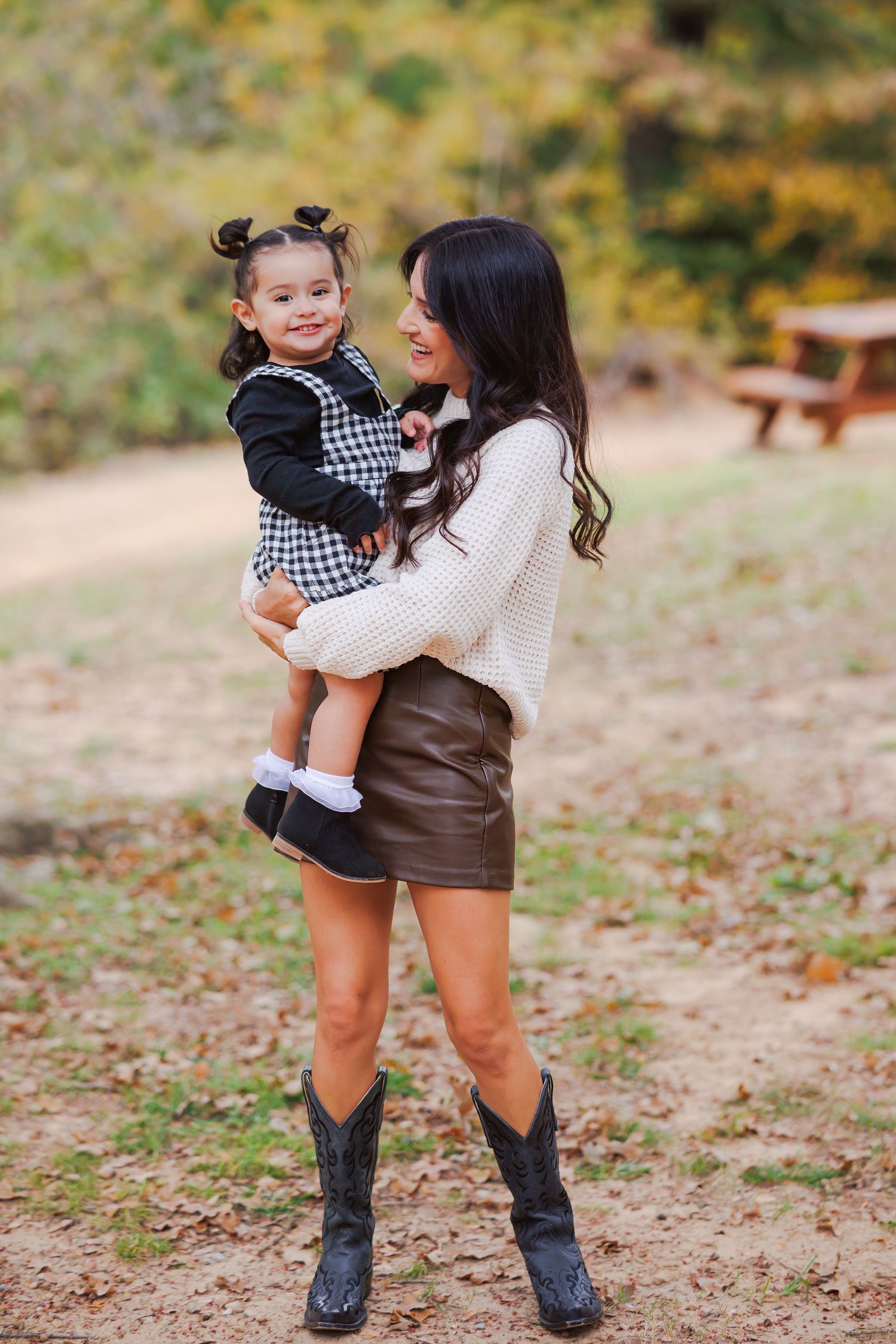 A mother and daughter in autumn setting wearing cozy sweaters and western boots, sharing a joyful outdoor moment at a park.