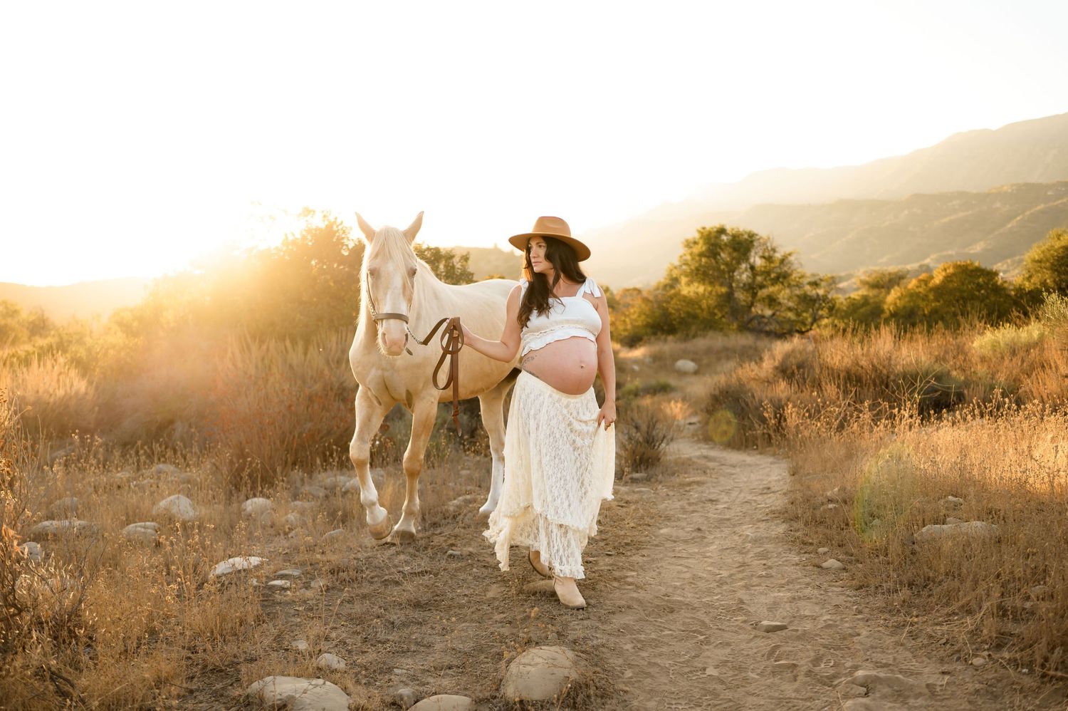 woman walks with white horse during sunset on trail at Ojai Valley Trail riding during her maternity photoshoot in Ojai while on babymoon