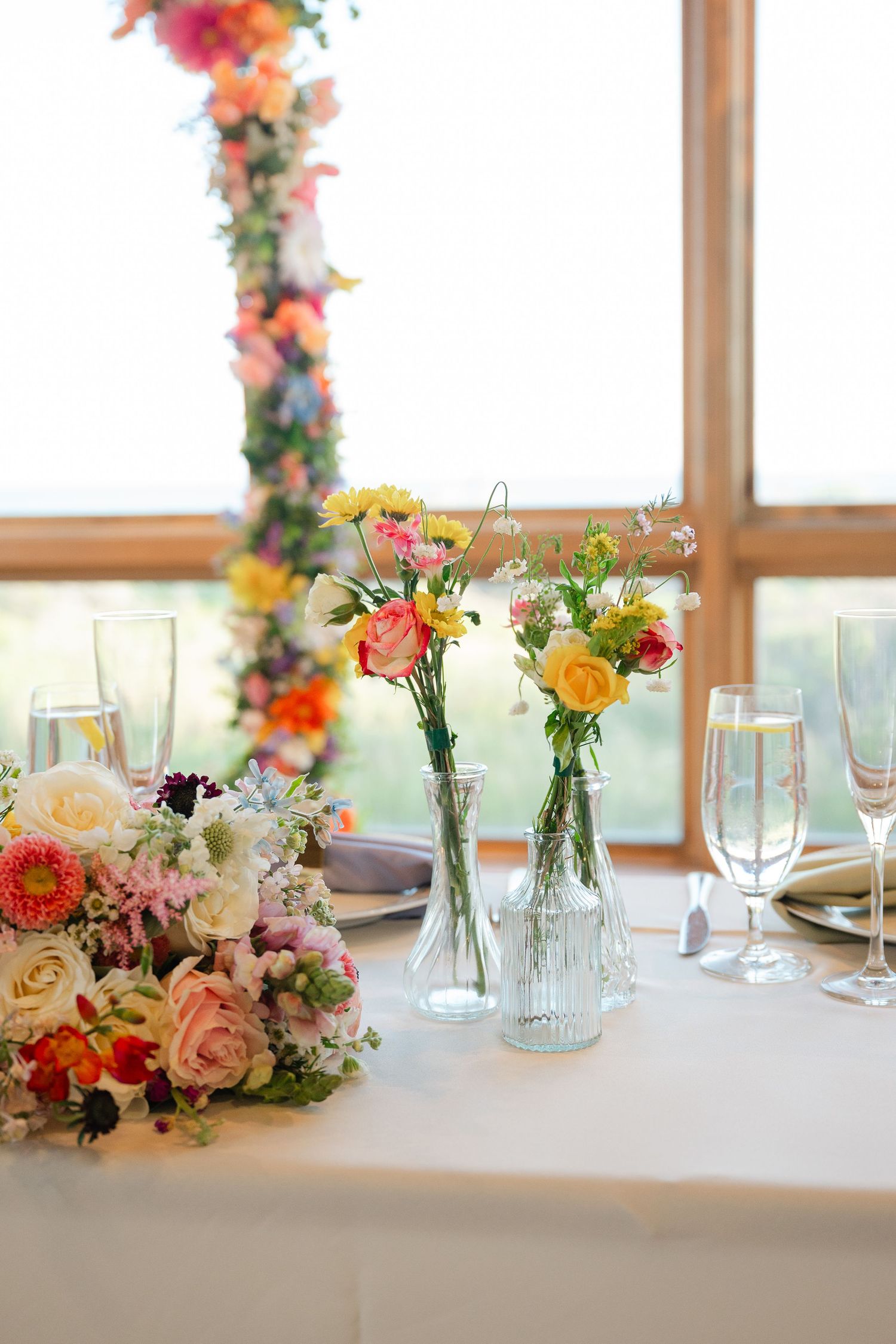 Elegant wedding reception table with colorful floral centerpieces in glass vases and champagne flutes on white tablecloth.
