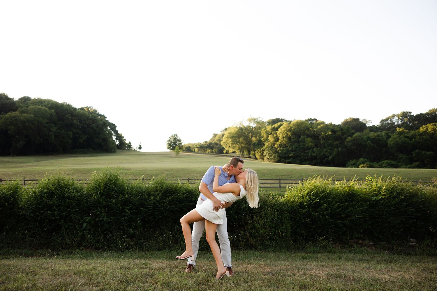 A couple embraces in a romantic dip pose at sunset in a grassy meadow with trees in the background.