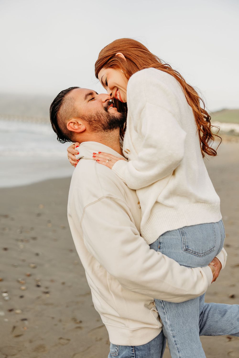 A romantic couple embracing and laughing together on a foggy beach wearing matching cream sweaters.