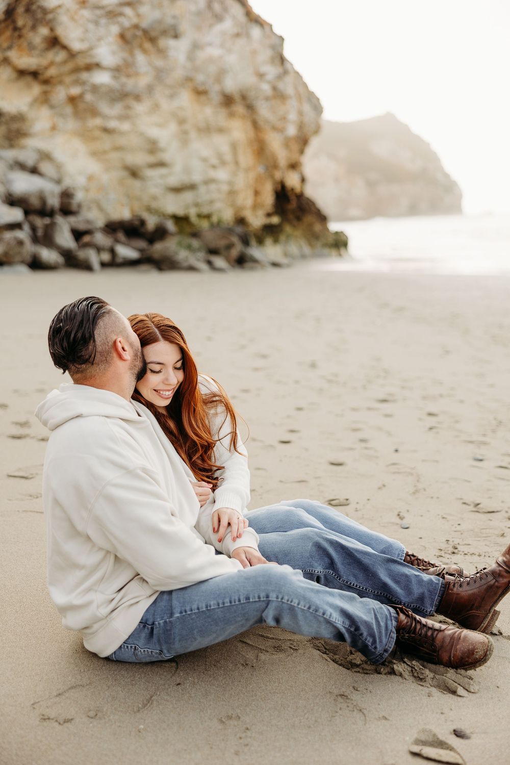 Couple in casual attire sits intimately on a foggy beach with dramatic coastal cliffs in the background.