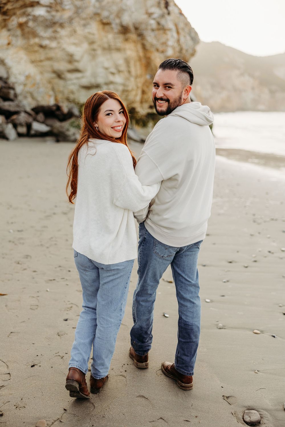 A couple wearing matching white sweaters and jeans walk together on a beach with cliffs in the background.