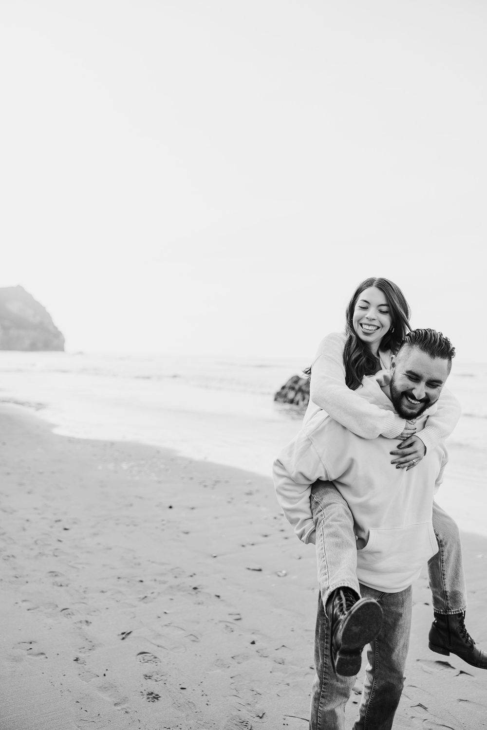 A joyful piggyback ride moment captured in black and white on a scenic beach with coastal cliffs in the background.