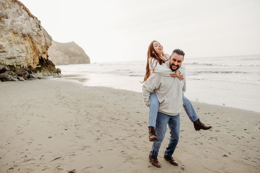 A couple playfully enjoying themselves on a beach with cliffs in the background during an overcast day.
