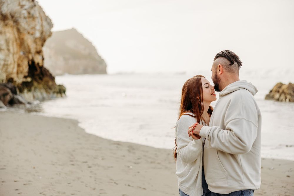 Loving couple sharing tender moments on a rocky beach with dramatic coastal cliffs in the background.