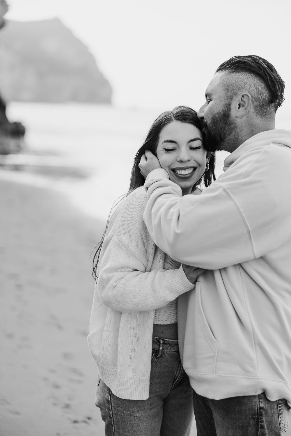 Black and white photo of a joyful couple embracing on a beach with cliffs in the background.