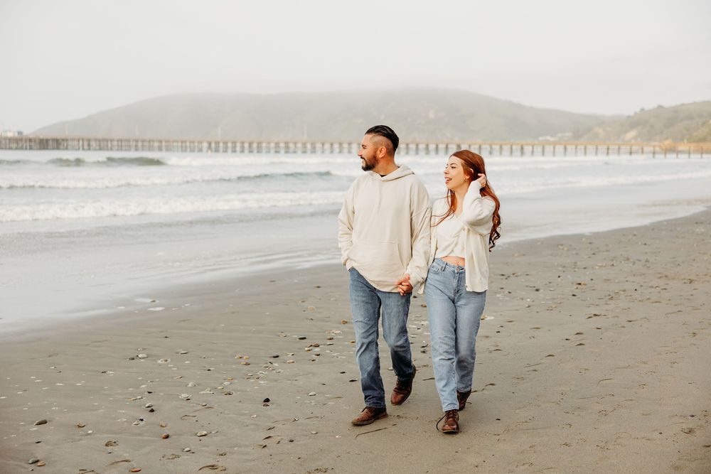 A couple walks hand in hand along a misty oceanfront beach wearing matching light denim and neutral colors.