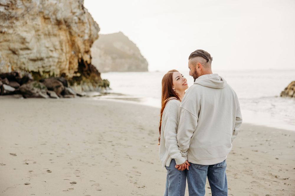A romantic couple sharing an intimate moment on a sandy beach with dramatic coastal rock formations behind them.