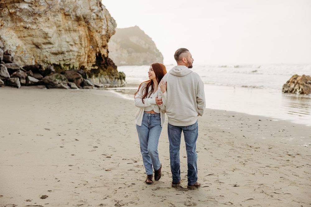 A couple walking hand in hand along a scenic beach with towering cliffs in the background.
