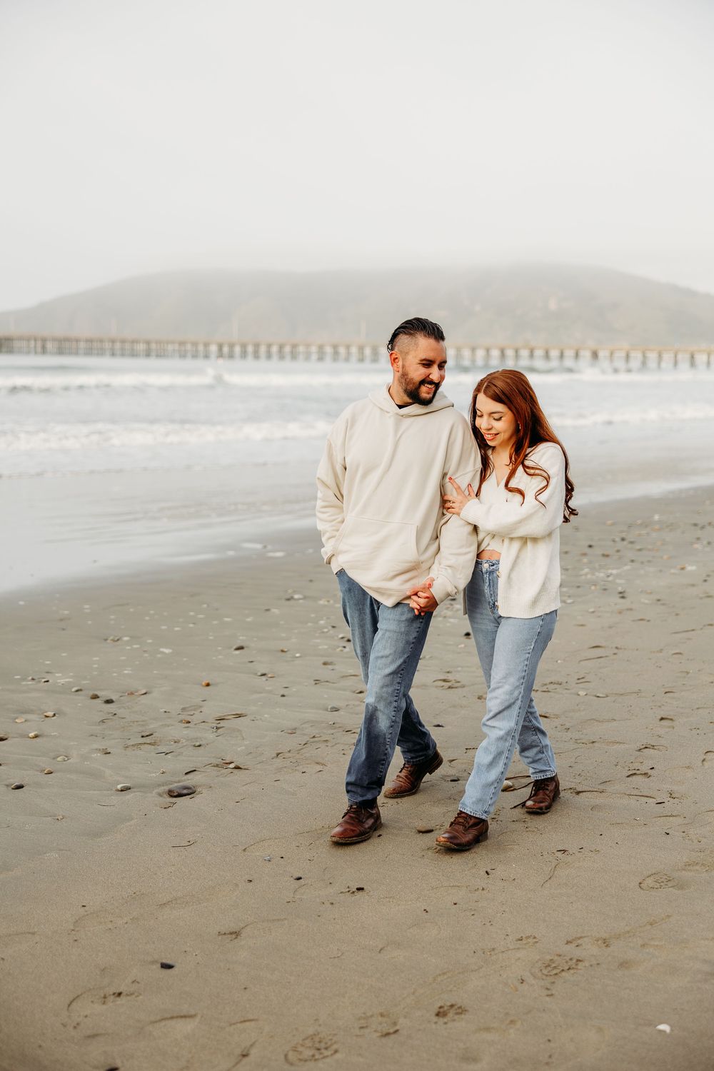 A couple walks together and laughs on a foggy beach with a pier visible in the background.