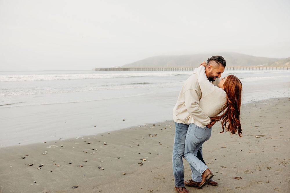 A couple shares an intimate moment on a misty beach with rolling waves and mountains in the background.