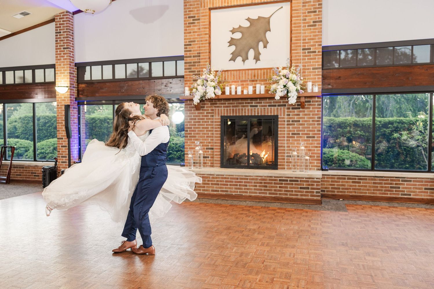 Dancing couple performing a dip move on wooden floor by brick fireplace at rustic reception venue.