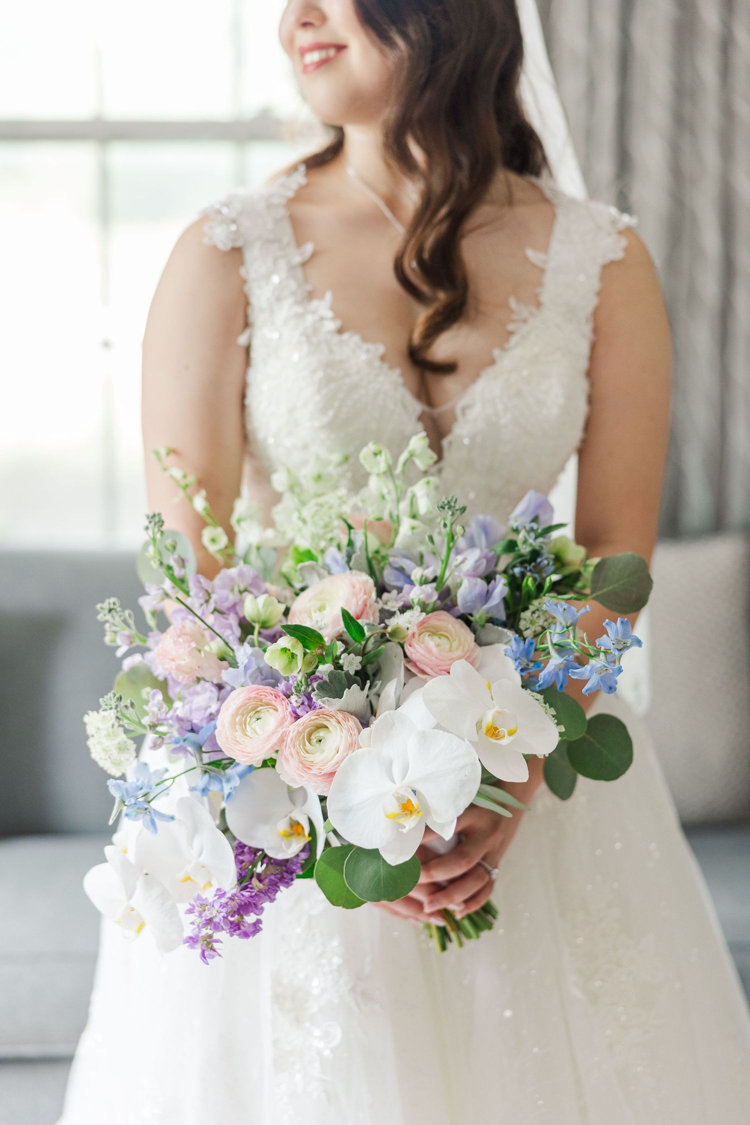 A bride in a lace wedding dress holds a romantic pastel bouquet with orchids, roses, and delicate blue flowers.