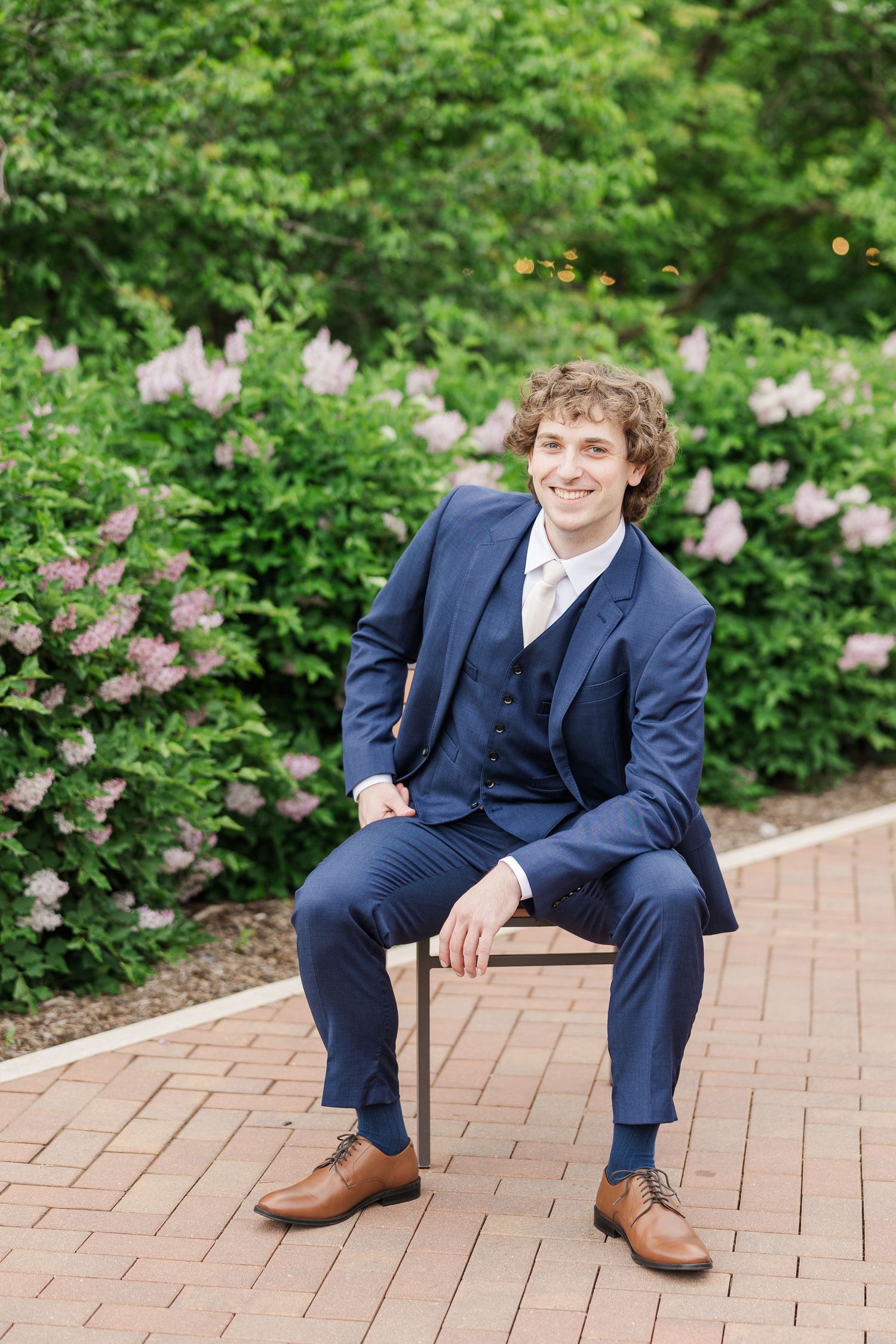 A person in a navy blue three-piece suit and brown shoes sits on a bench near blooming pink lilac bushes in a garden.