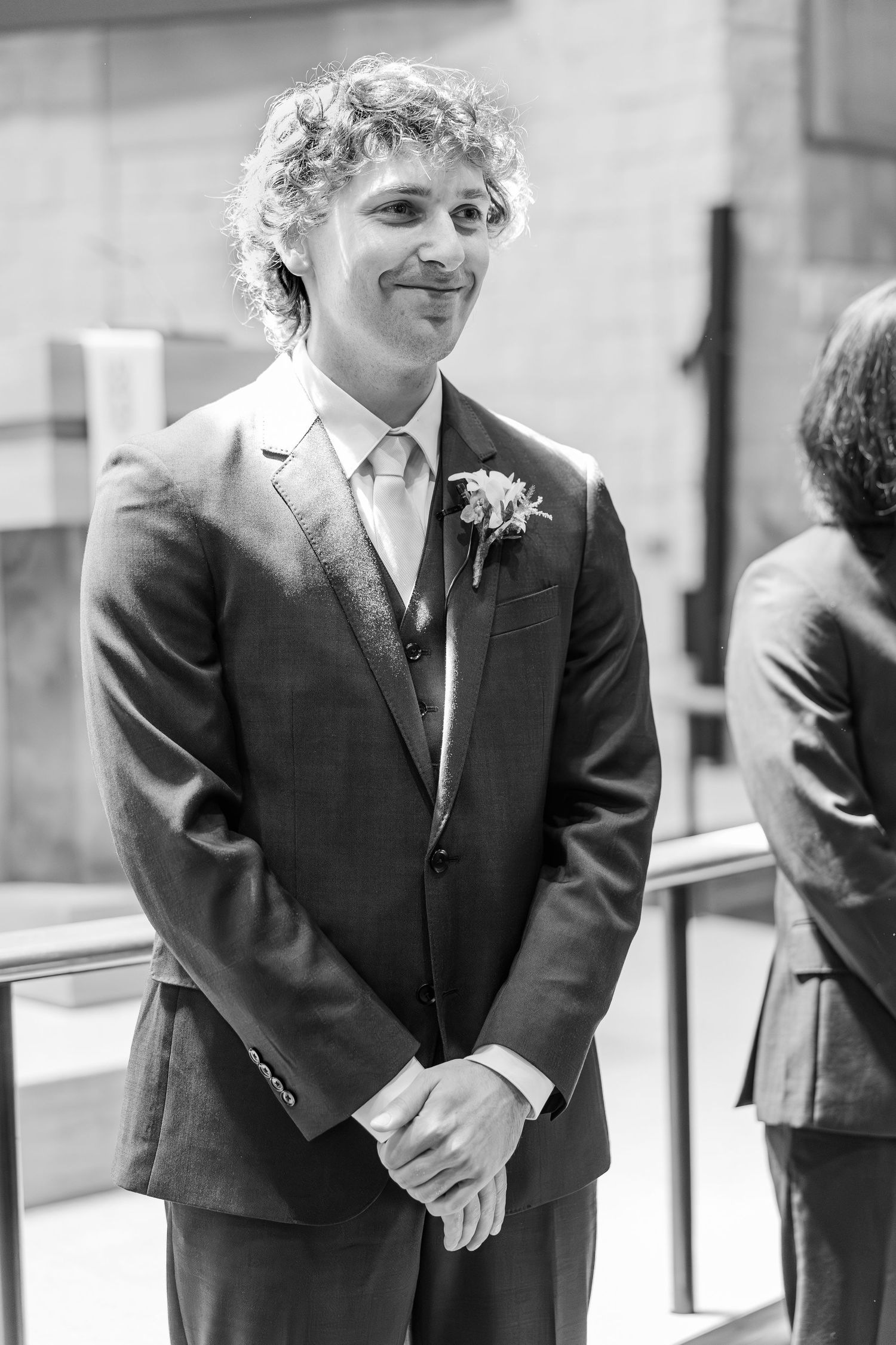 A groom in a dark suit with curly hair smiles during a wedding ceremony, wearing a boutonniere flower.