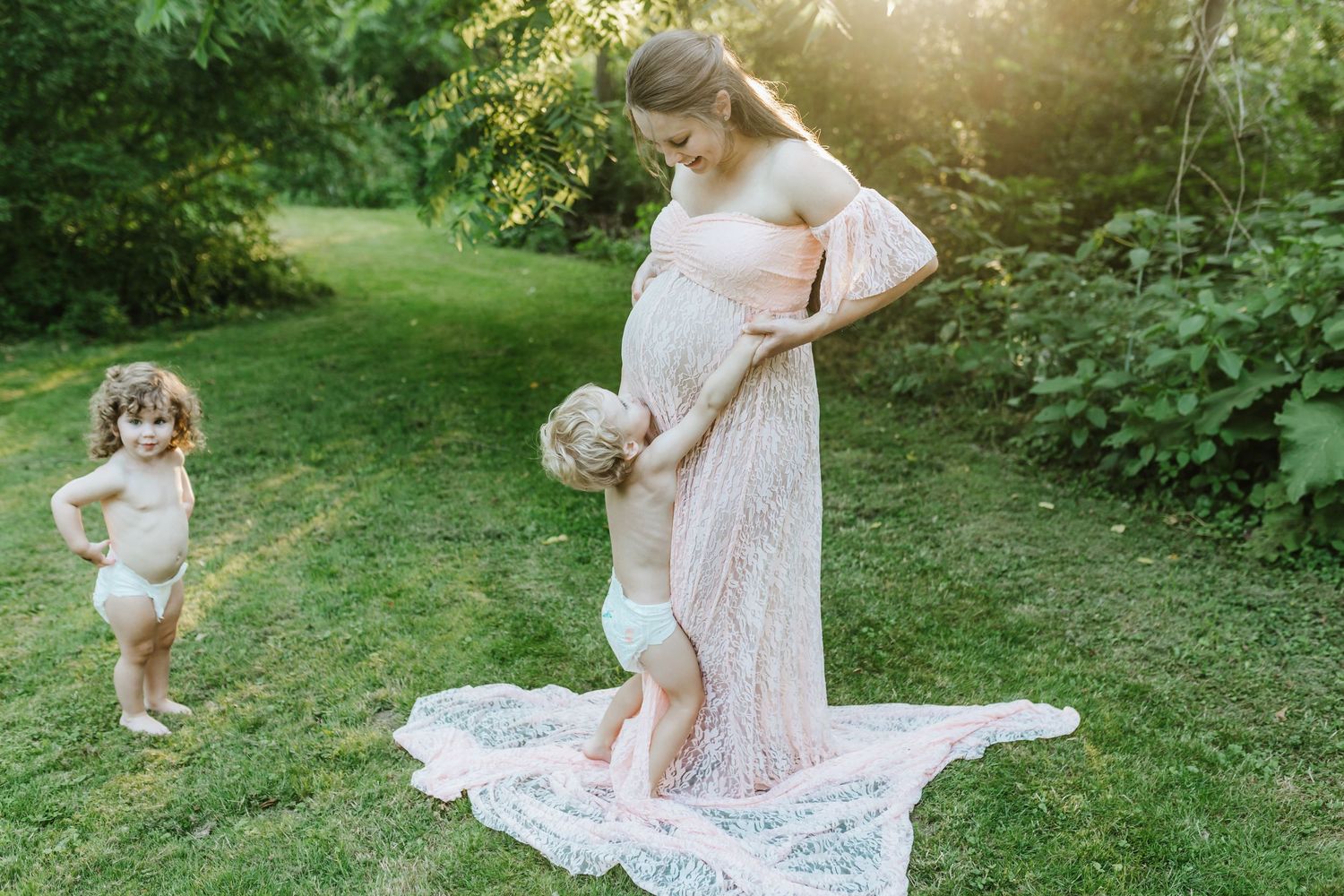A person in a lace dress stands in a sunlit garden while a young child plays nearby.
