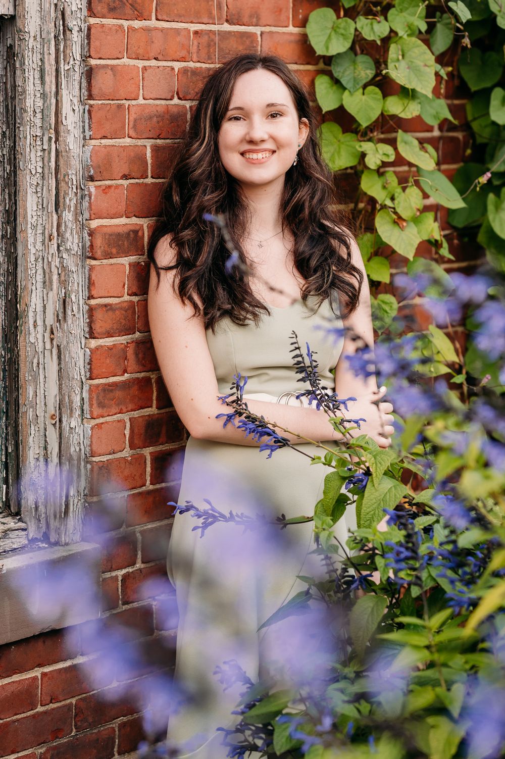 A person in a sage green dress poses against a brick wall with purple flowers in the garden foreground.