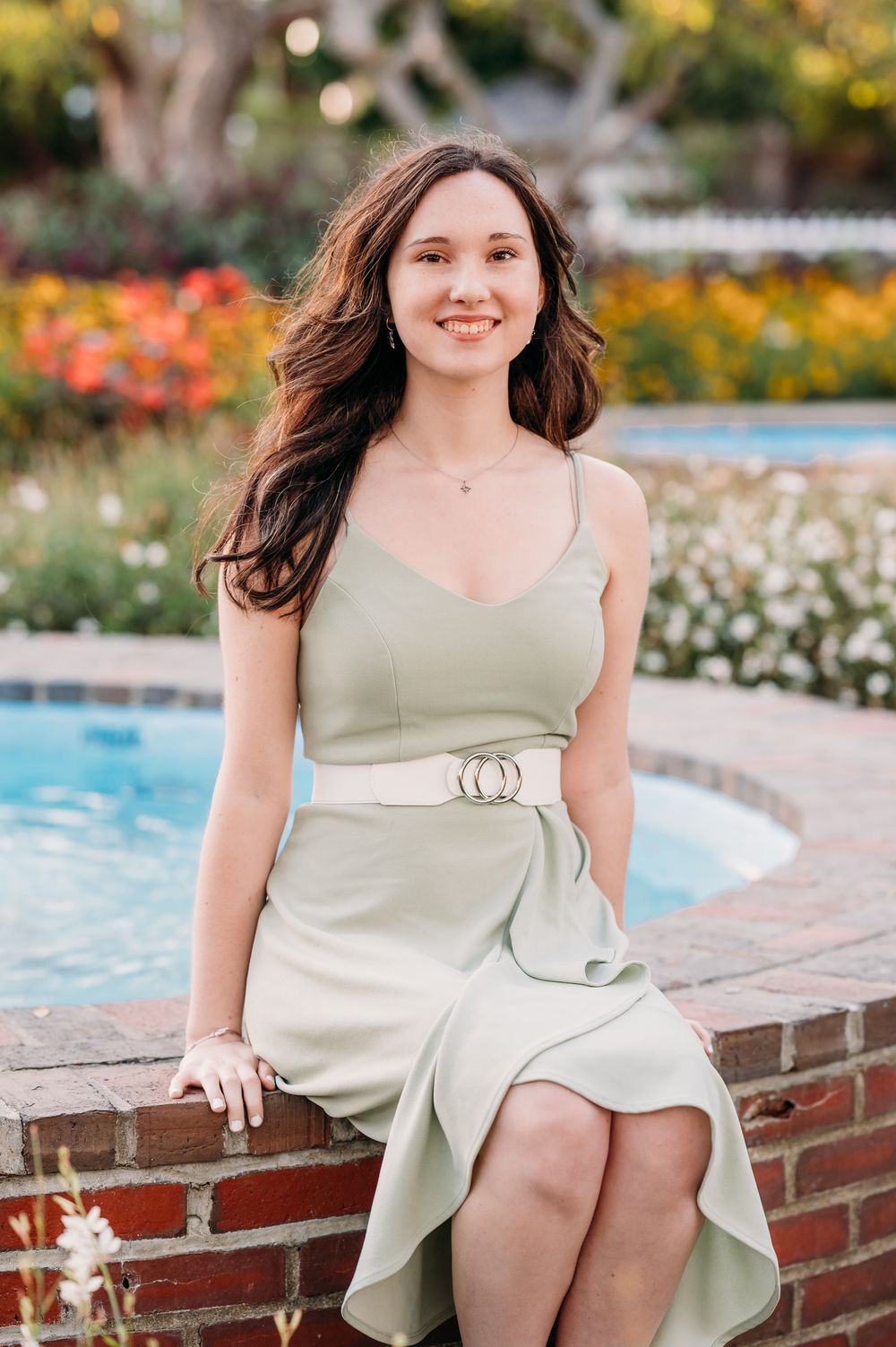 A woman in a sage green sundress with a statement belt sits elegantly by a poolside surrounded by colorful flowers.