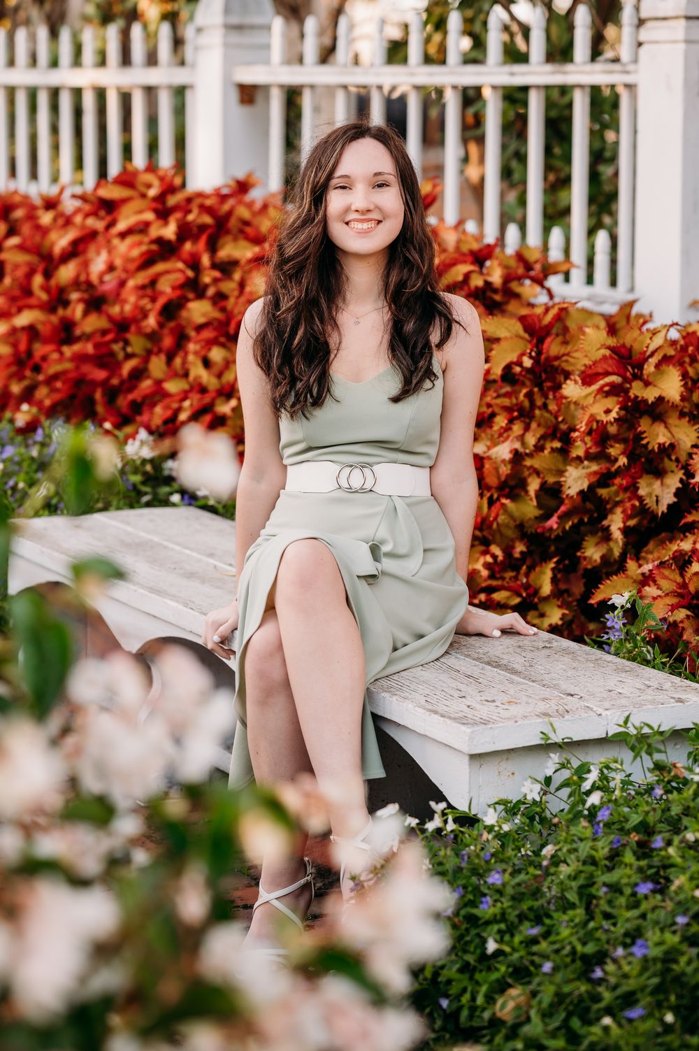A sage green dress and autumn foliage create a seasonal outdoor portrait on a white garden bench.
